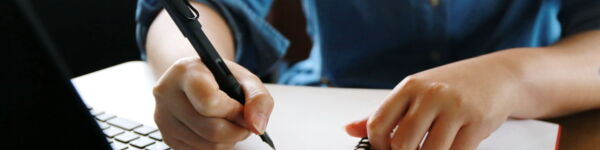 Image of a person sitting at a desk, writing in a notebook. A laptop and mobile phone is on the desk.